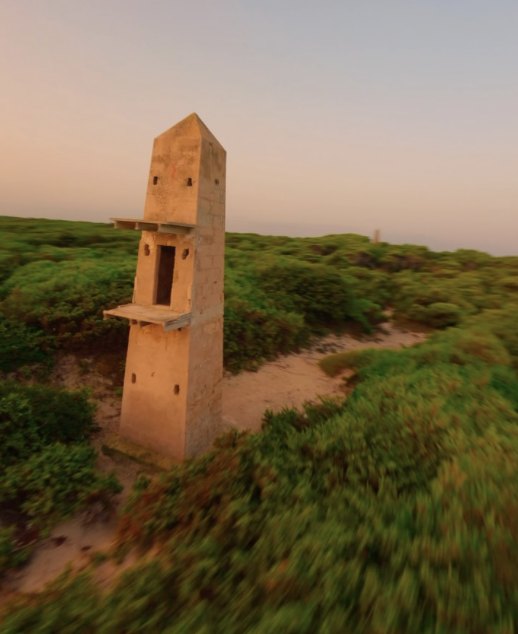 FPV drone flying low over beach dunes past an old concrete tower at sunset